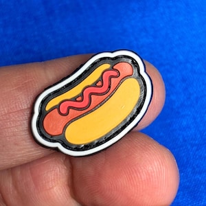 以下が含まれることがあります： 黒い縁取り、白い縁取り、明るい青色の背景が特徴的な、カラフルなホットドッグチャーム。 ホットドッグは、黄色のパン、赤いソーセージ、黄色のマスタードの波線が特徴です。