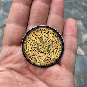 Puede incluir: Una pegatina circular negra y dorada con un diseño de estrella de siete puntas y los nombres de los ángeles alrededor del perímetro. Los nombres incluyen: Gabriel, Tzafqiel, Tzadkiel, Samael, Rafael, Haniel y Miguel.