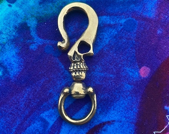 May include: A brass skull-shaped swivel hook with a ring on the bottom. The hook is a dark gold color and has a detailed skull design.