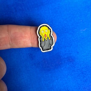 以下が含まれることがあります： エドヴァルド・ムンクの絵画「叫び」の漫画版をあしらった靴チャーム。チャームは黄色い顔、開いた口、頬に手を当てており、灰色の体と白い縁取りがあります。背景は青色です。