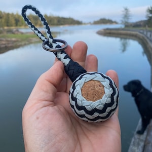 May include: A black and silver braided keychain with a cork centre. The keychain is held in a hand against a blurred background of water and trees.