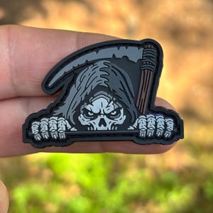 May include: A black, gray, and brown rubberized patch featuring a grim reaper character with a hooded skull face, skeletal hands gripping a horizontal bar, and a scythe blade positioned behind the head against a solid black background.