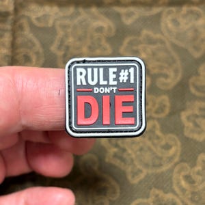 Könnte beinhalten: Schwarz-weißes Patch mit weißem Rand. Das Patch hat den Text "RULE #1 - DON'T - DIE" in weißen und roten Buchstaben.