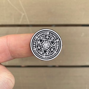 May include: A black and white round pin featuring a detailed esoteric design. The pin has a circular border with text and symbols, surrounding a central star-like pattern. The design includes crosses, moons, and other geometric shapes.