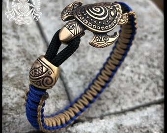 May include: A blue and gold paracord bracelet with a bronze Maori sea turtle charm. The charm features intricate carvings and a spiral design. The bracelet is adjustable and has a secure clasp.