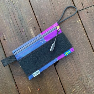 Può includere: Una custodia rettangolare con un design sfumato viola e blu, una toppa in velcro nera e una cerniera dorata. La custodia ha un anello nero e una piccola etichetta con la scritta "KRUGER". È visibile una piccola bandiera svedese.
