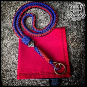 Op de afbeelding: Een lanyard gemaakt van gevlochten blauw en rood koord met een zilverkleurige metalen kraal met een gestileerde letter S, een bijpassend gevlochten handvat en een ronde messingkleurige metalen ring aan een vierkant rood stoffen tasje.
