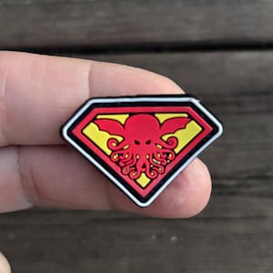 May include: A red and yellow superhero patch featuring a Cthulhu-like creature in the centre. The patch is diamond-shaped and has a white border.