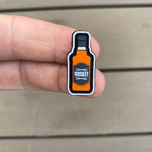 以下が含まれることがあります： 「WHISKEY」と書いてある白いラベルが付いた、黒とオレンジのゴム製のウイスキーボトルの形をしたチャームです。