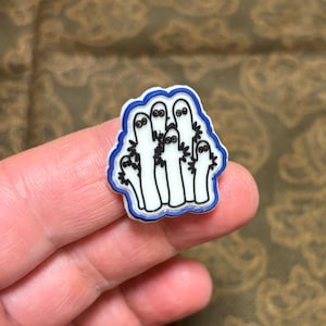 May include: A white enamel pin with a blue outline depicting a group of cartoon ghosts with black eyes and mouths. The ghosts are arranged in a cluster, with some standing taller than others.