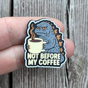 Może przedstawiać: Zawieszka do butów przedstawiająca postać z kreskówki Godzilli trzymającą filiżankę kawy. Zawieszka jest szara, z pomarańczowymi i czarnymi akcentami oraz białym napisem "NOT BEFORE MY COFFEE".