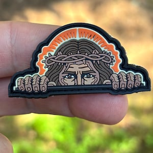 Könnte beinhalten: Ein gummiertes Abzeichen mit einer grafischen Illustration einer Figur mit langem braunem Haar, die eine Dornenkrone trägt. Die Figur ist hinter einem schwarzen horizontalen Balken positioniert, mit einem orangefarbenen Heiligenschein, schwarz umrandet.