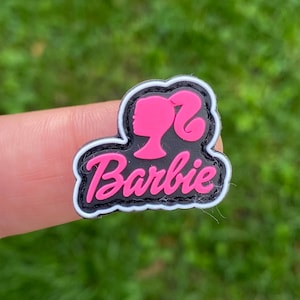 Puede incluir: Un parche de goma rosa y negro con un contorno blanco. El parche presenta el logotipo de Barbie con una silueta rosa de la cabeza de una mujer y la palabra "Barbie" en letras rosas.