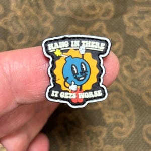 Può includere: Spilla colorata con un personaggio a forma di bomba dei cartoni animati. La bomba ha una faccia blu, un corpo giallo e una miccia rossa. La spilla recita "HANG IN THERE" sopra la bomba e "IT GETS WORSE" sotto.