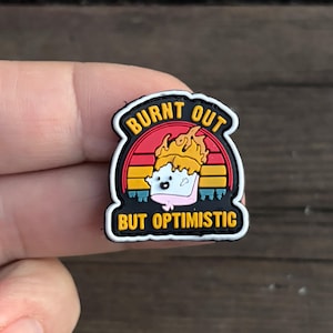 Könnte beinhalten: Ein schwarz-weißes Patch mit einem Cartoon-Bild eines verbrannten Marshmallows mit dem Text "Burnt Out But Optimistic".