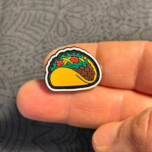 Puede incluir: Un colorido dije de taco con tortilla amarilla, carne marrón, lechuga verde y tomates rojos. El dije tiene un borde blanco y negro. Este accesorio temático de comida es perfecto para añadir un toque divertido a zapatos u otros artículos.
