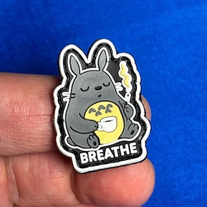 Könnte beinhalten: Ein grauer Cartoon-Kaninchen-Charakter mit gelbem Bauch und weißer Tasse, der eine brennende Zigarette hält. Das Wort "BREATHE" ist unten in Weiß gedruckt. Das Bild ist ein Schuhanhänger.