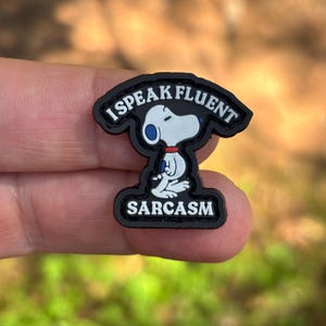 May include: A black shoe charm featuring a white cartoon dog with a red collar and blue ear detail. The text I SPEAK FLUENT is arched above the dog, and the word SARCASM is printed in white capital letters at the bottom of the black base.