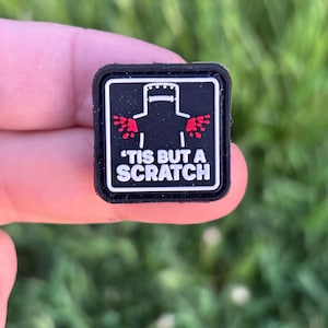 May include: A black square patch with a white border features a stylized illustration of a knight with red accents and the text "'TIS BUT A SCRATCH" in white. The patch is held in a person's hand.