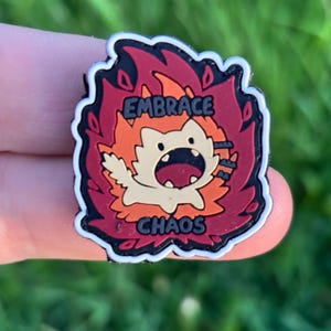 Peut inclure: Une épingle colorée représentant un personnage de dessin animé en flammes. L'épingle porte les mots "EMBRACE" et "CHAOS" en texte blanc. Le personnage est orange et blanc, entouré de flammes rouges et oranges, avec une bordure noire.