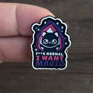 May include: Black cat wearing a pink and purple hooded cloak with stars and moons. The text reads "F**k Normal I Want Magic".