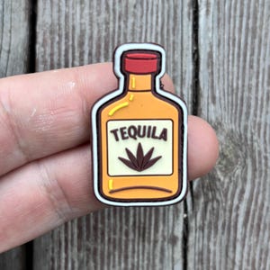 Puede incluir: Un colorido dije con forma de botella de tequila. La botella es naranja con una tapa roja y una etiqueta blanca que dice "TEQUILA" con una ilustración de agave marrón. El dije tiene un contorno marrón.