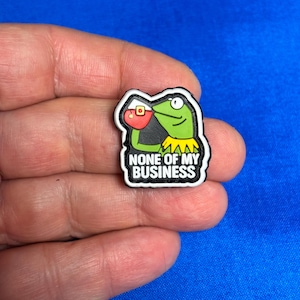 以下が含まれることがあります： 赤い飲み物を持っている緑のカエルのキャラクターが特徴の靴チャーム。チャームは黒い縁取りで、「NONE OF MY BUSINESS」というテキストが白で表示されています。背景は無地の青色です。