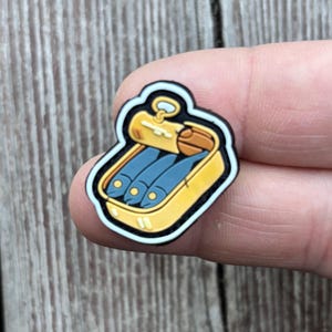 以下が含まれることがあります： 3匹の青い魚が入った黄色のイワシ缶のカラフルなチャーム。缶には茶色の蓋とプルタブが付いています。チャームは白と黒で縁取られています。