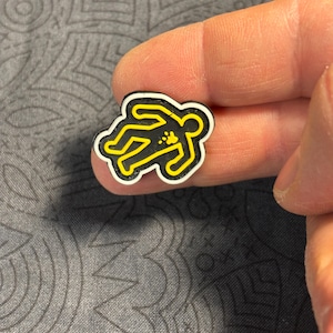 May include: A small, black and yellow pin shaped like a chalk outline of a body at a crime scene. The pin has a white border and is held between fingers against a gray patterned background.