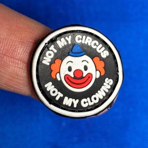 Puede incluir: Un dije redondo negro con el texto blanco "NOT MY CIRCUS NOT MY CLOWNS". El dije presenta un payaso de dibujos animados con pelo naranja, nariz y boca rojas y un sombrero azul. El dije está sujeto por un dedo.