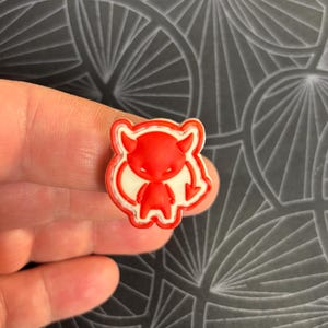 May include: A small red devil figurine with white outlines. The devil has horns, a tail, and a mischievous expression.