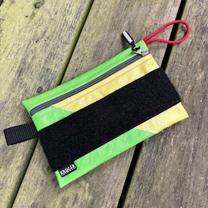 May include: A small, rectangular pouch with a lime green and yellow design, a black velcro strap, and a black zipper. The pouch has a red loop and a black strap. The word "KRUGER" is printed on the bottom corner.