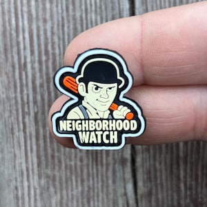 Könnte beinhalten: Ein Schuhanhänger mit einer Cartoonfigur in einem schwarzen Melone, die einen Baseballschläger hält. Der Anhänger ist schwarz, weiß, orange und beige. Der Text "NEIGHBORHOOD WATCH" ist weiß auf schwarzem Hintergrund.