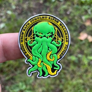 Peut inclure: Un patch circulaire vert et jaune avec une image noire et blanche d'une créature tentaculaire. Le texte autour de la créature dit "Iä! Iä! Cthulhu Fhtagn!"