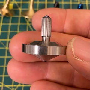 Op de afbeelding: Een zilveren metalen draaitop met een geribbelde, zilveren metalen steel. De draaitop wordt in de hand van een persoon gehouden.