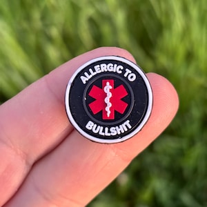 Könnte beinhalten: Ein runder, schwarz-weißer Charm mit dem weißen Schriftzug "ALLERGIC TO BULLSHIT". In der Mitte befindet sich ein rotes Medizinsymbol mit weißem Stab.