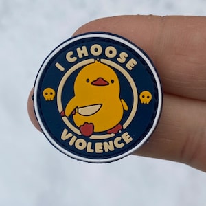 Puede incluir: Un parche redondo azul y blanco con un pato amarillo de dibujos animados sosteniendo un cuchillo. El texto "I CHOOSE VIOLENCE" está escrito en un círculo blanco alrededor del pato. Hay dos calaveras y huesos cruzados a cada lado del pato.