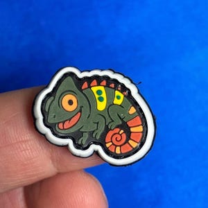 May include: A colorful chameleon-shaped charm with a white border. The charm features a green body, yellow and orange accents, and an orange eye. The chameleon is smiling, and the background is a solid blue.