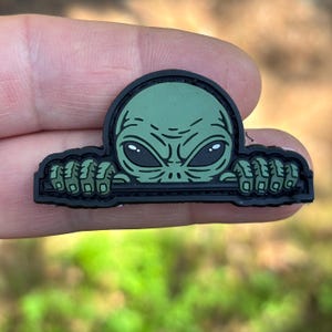 May include: A rubberized patch featuring a green alien head with large black eyes and textured hands gripping a horizontal black bar. The design is outlined in a thick black border, creating a distinct graphic element with a matte finish.