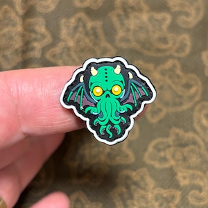 May include: A green and black Cthulhu patch with yellow eyes and tentacles. The patch is surrounded by a white border.