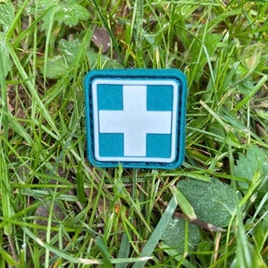 Puede incluir: Un parche médico azul verdoso con una cruz blanca en el centro. El parche está rodeado por un borde negro y tiene una superficie texturizada.