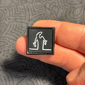 Può includere: Una piccola toppa quadrata nera con un disegno a tratto bianco di una persona con cappello e bastone. La figura è in piedi su una linea bianca. La toppa è tenuta da una mano.