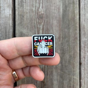 May include: Black square patch with a white middle finger and the words "FUCK CANCER" in red and yellow letters.