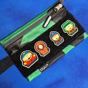 以下が含まれることがあります： 黒と緑の迷彩柄のポーチで、ファスナー開閉式です。ポーチには、4つの漫画のキャラクターパッチと「KRUGER」の文字がプリントされた黒いベルクロストリップが付いています。ファスナーの引き手には「SCUZZYISM」の文字がプリントされています。