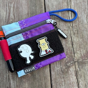 May include: A purple and blue zippered pouch with a black strap and two patches. The patches are of a cartoon dog and a cartoon man with glasses. The pouch has the word "KRUGGER" printed on it.