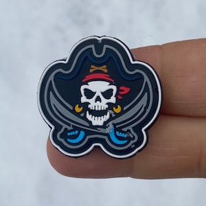 Peut inclure: Un crâne de pirate noir et blanc avec un bandana rouge et un chapeau noir, avec des épées croisées derrière le crâne. L'image est sur un fond noir et gris.