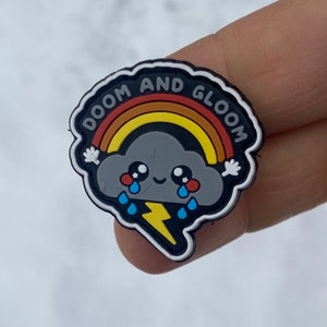 Peut inclure: Un charm de chaussure noir et blanc avec un nuage de dessin animé. Le nuage est gris avec un arc-en-ciel, des larmes et un éclair. Le texte "DOOM AND GLOOM" est écrit sur le bord du charm.