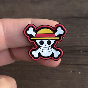 May include: A red, yellow, and white rubber charm with a skull and crossbones design. The skull is wearing a red and yellow straw hat.