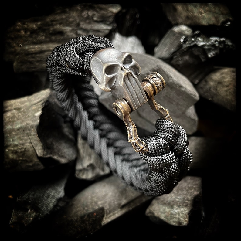 May include: A black paracord bracelet with a skull-shaped metal clasp. The clasp is made of brass and has a detailed skull design.