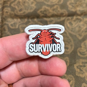 Könnte beinhalten: Weißer Patch mit einer roten Kakerlaken-Karikatur und dem Wort "SURVIVOR" in schwarzen Buchstaben.
